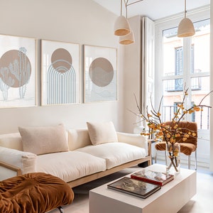 May include: A white sofa with two pillows sits in a living room with a white coffee table and a brown chair. Three framed prints with abstract designs hang on the wall behind the sofa. A branch with yellow berries is in a vase on the coffee table.