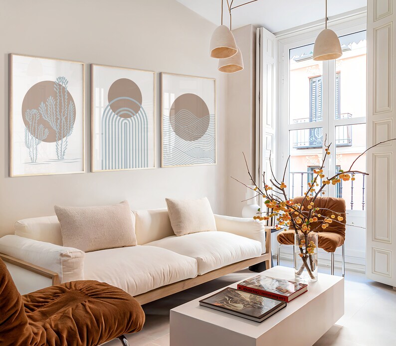May include: A white sofa with two pillows sits in a living room with a white coffee table and a brown chair. Three framed prints with abstract designs hang on the wall behind the sofa. A branch with yellow berries is in a vase on the coffee table.