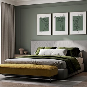 May include: A bedroom with a grey bed, green bedding, and a yellow bench at the foot of the bed. There are three framed prints on the wall behind the bed, each with a line drawing of a plant. A small bedside table with two drawers is on the right side of the bed.
