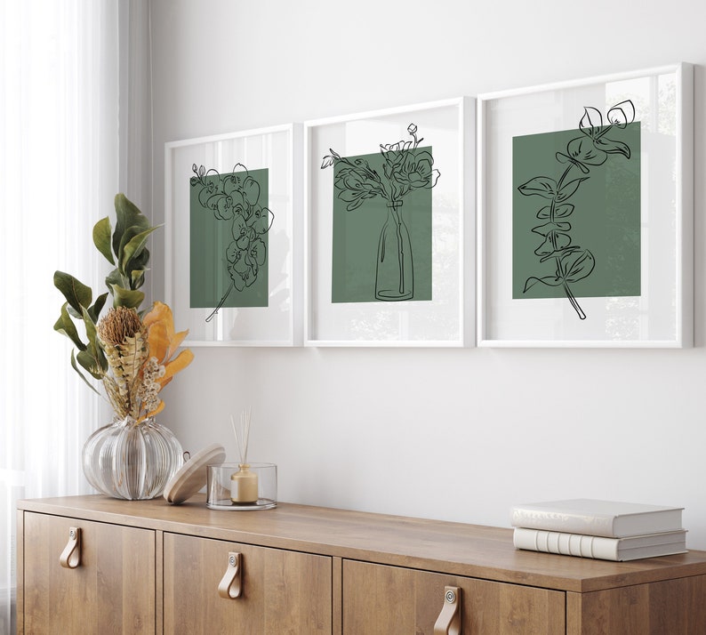 May include: Three white framed prints featuring black line drawings of flowers and foliage on a green background. The prints are displayed on a white wall above a wooden cabinet.