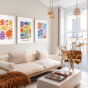 K&ouml;nnte beinhalten: Drei gerahmte Blumenprints mit bunten Blumen auf wei&szlig;em Hintergrund h&auml;ngen an einer hellbeigen Wand &uuml;ber einem wei&szlig;en Sofa. Die Drucke sind mit "Flower Market" und "You Are My Sunshine" betitelt.