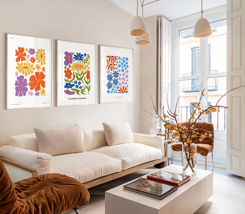 K&ouml;nnte beinhalten: Drei gerahmte Blumenprints mit bunten Blumen auf wei&szlig;em Hintergrund h&auml;ngen an einer hellbeigen Wand &uuml;ber einem wei&szlig;en Sofa. Die Drucke sind mit "Flower Market" und "You Are My Sunshine" betitelt.
