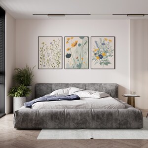 May include: Three framed floral art prints hang above a gray upholstered bed. The prints feature botanical illustrations in shades of green, yellow, and blue. A large potted plant sits to the left of the bed, and a small side table is on the right.