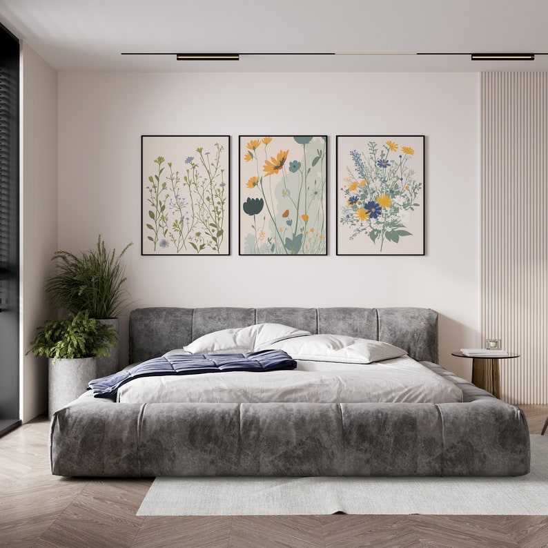 May include: Three framed floral art prints hang above a gray upholstered bed. The prints feature botanical illustrations in shades of green, yellow, and blue. A large potted plant sits to the left of the bed, and a small side table is on the right.
