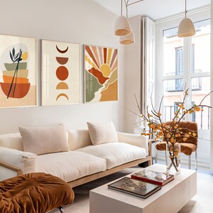 May include: A living room interior with a white sofa, a brown armchair, and a white coffee table. Three abstract art prints with geometric shapes and colours hang on the wall above the sofa. A vase with yellow flowers sits on the coffee table.