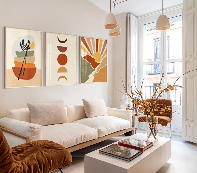 May include: A living room interior with a white sofa, a brown armchair, and a white coffee table. Three abstract art prints with geometric shapes and colours hang on the wall above the sofa. A vase with yellow flowers sits on the coffee table.