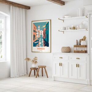 May include: A white wall with a gold framed poster of a colorful illustration of Venice, Italy. The poster has the text "Venice, Italy" on it. The wall is decorated with two gold shelves with various items on them. There is a white cabinet with gold handles below the shelves.
