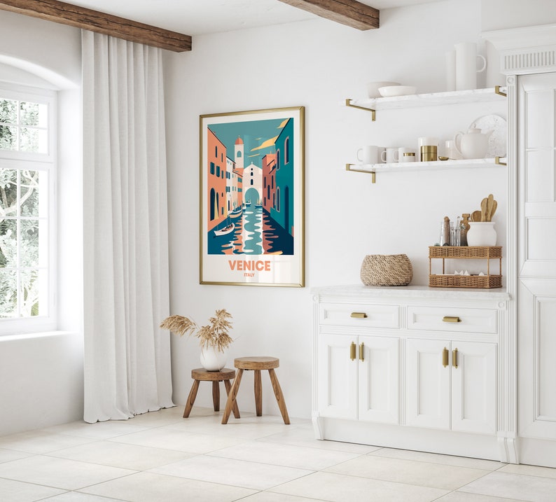 May include: A white wall with a gold framed poster of a colorful illustration of Venice, Italy. The poster has the text "Venice, Italy" on it. The wall is decorated with two gold shelves with various items on them. There is a white cabinet with gold handles below the shelves.