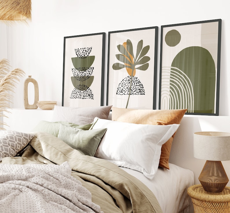 May include: Three abstract art prints in black frames with a white background. The prints feature green, brown, and white geometric shapes and patterns. The prints are hanging on a white wall above a bed with white and green bedding.