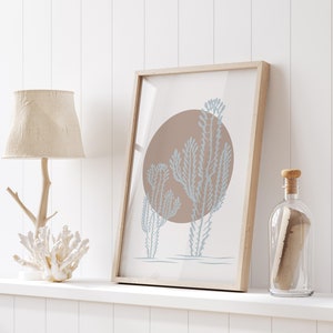 May include: A framed print with a minimalist design featuring three blue-grey stylized plants against a brown circle on a white background. The print is displayed on a white shelf with a wooden lamp and a clear glass bottle.