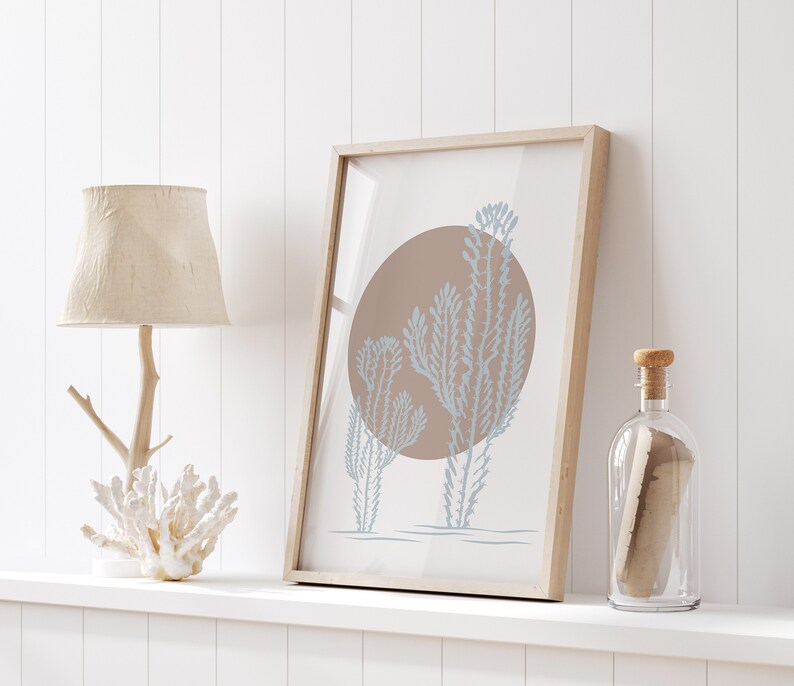 May include: A framed print with a minimalist design featuring three blue-grey stylized plants against a brown circle on a white background. The print is displayed on a white shelf with a wooden lamp and a clear glass bottle.