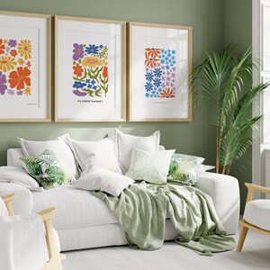 K&ouml;nnte beinhalten: Drei gerahmte Blumenprints mit einem wei&szlig;en Sofa und einer gr&uuml;nen Wolldecke in einem Wohnzimmer. Die Drucke zeigen bunte Blumen im Retro-Stil.