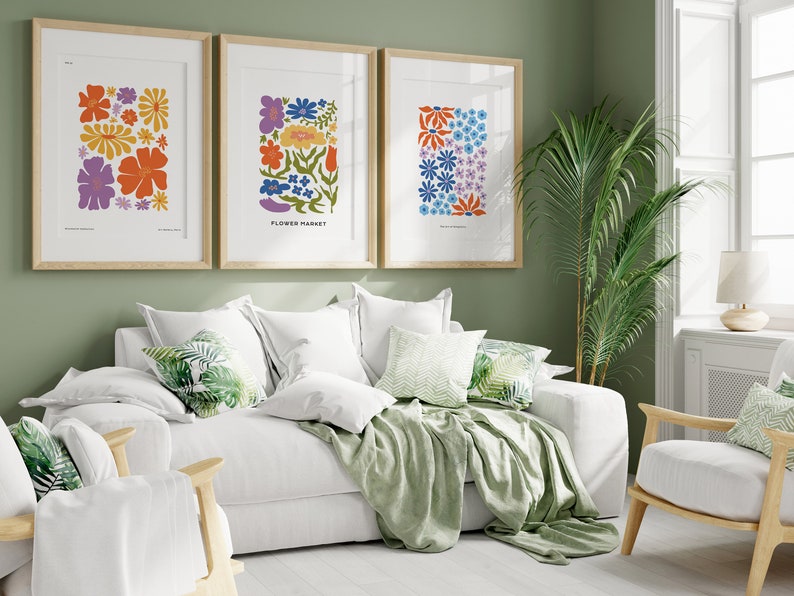 K&ouml;nnte beinhalten: Drei gerahmte Blumenprints mit einem wei&szlig;en Sofa und einer gr&uuml;nen Wolldecke in einem Wohnzimmer. Die Drucke zeigen bunte Blumen im Retro-Stil.
