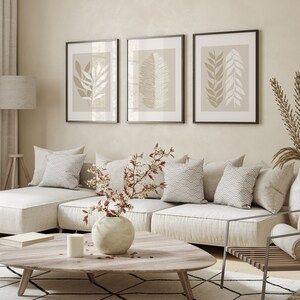 May include: A white sectional sofa with beige cushions and throw pillows, a wooden coffee table with a vase of white flowers, and three framed prints with beige and white botanical illustrations on a light beige wall.