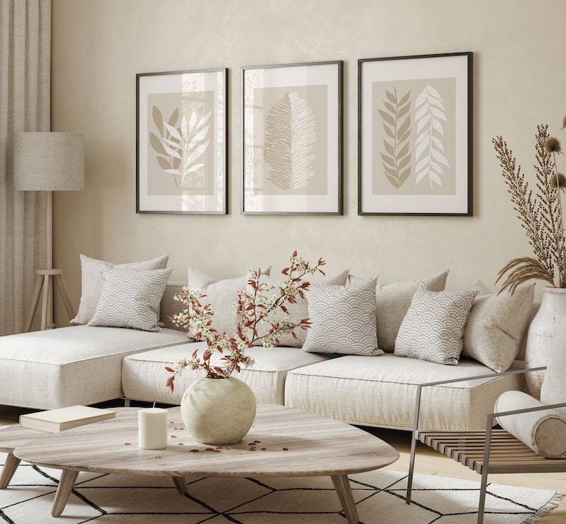 May include: A white sectional sofa with beige cushions and throw pillows, a wooden coffee table with a vase of white flowers, and three framed prints with beige and white botanical illustrations on a light beige wall.
