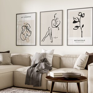 May include: Three black and white minimalist art prints with beige backgrounds. The prints are framed in black frames and hang on a white wall above a beige sectional sofa. The prints feature abstract line drawings of a figure, a plant, and a swirl.