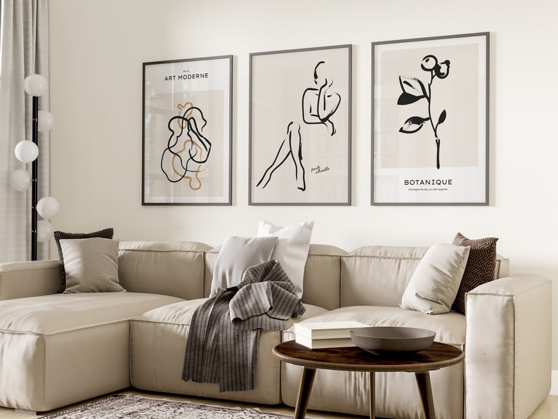 May include: Three black and white minimalist art prints with beige backgrounds. The prints are framed in black frames and hang on a white wall above a beige sectional sofa. The prints feature abstract line drawings of a figure, a plant, and a swirl.