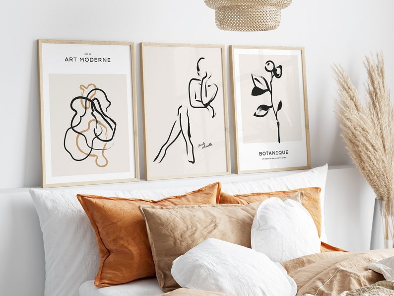 May include: Three framed prints with abstract line drawings in black and white. The prints are hung on a white wall above a bed with brown and white pillows. The prints are titled "Art Moderne", "Brush Strokes", and "Botanique".