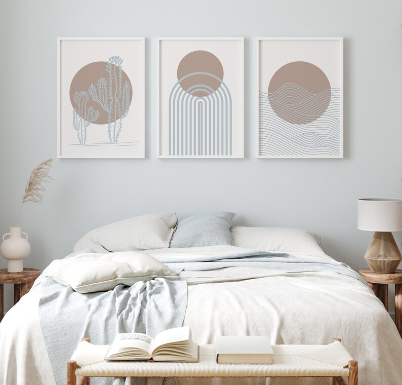 May include: Three framed abstract prints with a minimalist design. The prints feature a brown sun with blue and white geometric shapes. The prints are hung on a light blue wall above a white bed with a white blanket and pillows.