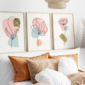 May include: Three framed prints with abstract line drawings of flowers in pink, blue, and green colors. The prints are hung on a white wall above a bed with brown and white pillows.