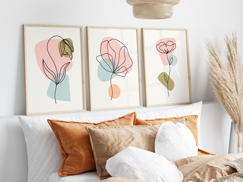 May include: Three framed prints with abstract line drawings of flowers in pink, blue, and green colors. The prints are hung on a white wall above a bed with brown and white pillows.
