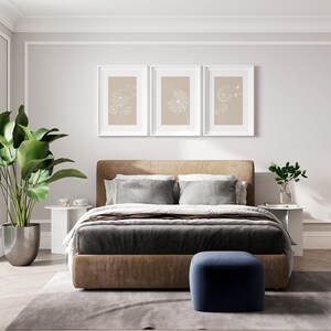 May include: A bedroom with a brown upholstered bed with a gray duvet and white pillows. There are three framed prints above the bed with a beige background and white dandelion designs. A small potted plant is on a white side table next to the bed. A blue ottoman sits in front of the bed on a gray rug.