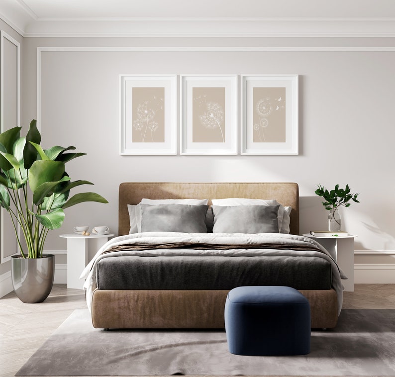 May include: A bedroom with a brown upholstered bed with a gray duvet and white pillows. There are three framed prints above the bed with a beige background and white dandelion designs. A small potted plant is on a white side table next to the bed. A blue ottoman sits in front of the bed on a gray rug.