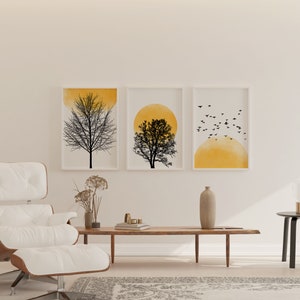 May include: Three framed prints with a minimalist design featuring a silhouette of a tree against a yellow watercolor background. The prints are hung on a white wall above a white leather armchair and a wooden coffee table.