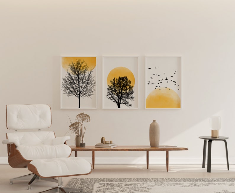 May include: Three framed prints with a minimalist design featuring a silhouette of a tree against a yellow watercolor background. The prints are hung on a white wall above a white leather armchair and a wooden coffee table.