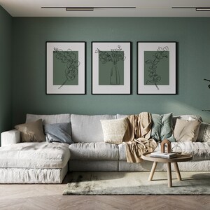 May include: A white sectional sofa with cushions and a throw blanket sits in front of a sage green wall with three black framed prints of line drawings of flowers. A wooden coffee table with a book and decorative objects sits in front of the sofa.