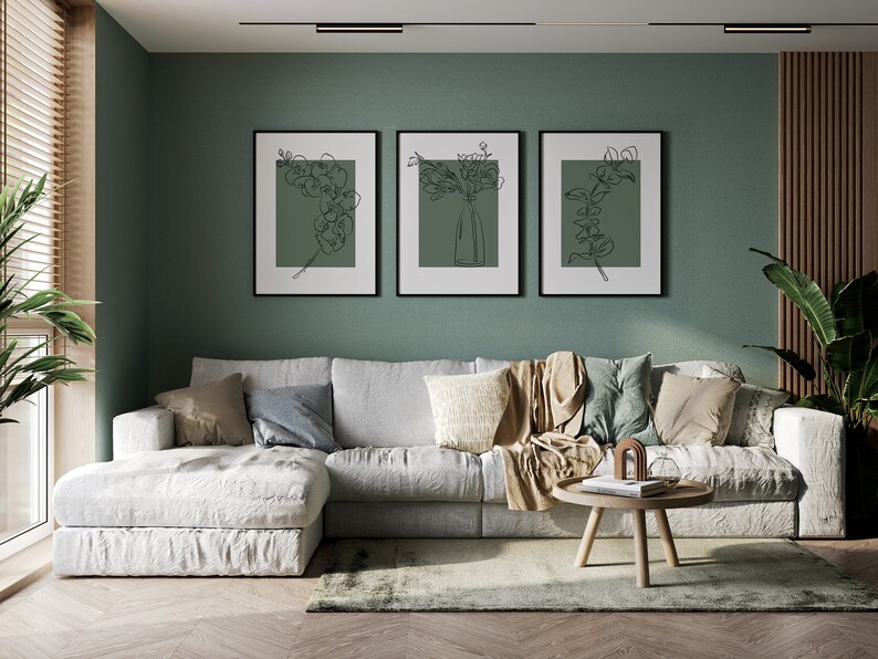 May include: A white sectional sofa with cushions and a throw blanket sits in front of a sage green wall with three black framed prints of line drawings of flowers. A wooden coffee table with a book and decorative objects sits in front of the sofa.