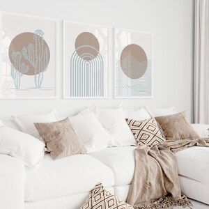 May include: Three abstract art prints with a minimalist design featuring a brown circle and blue lines. The prints are framed in white and hung on a white wall above a white couch with brown throw pillows.