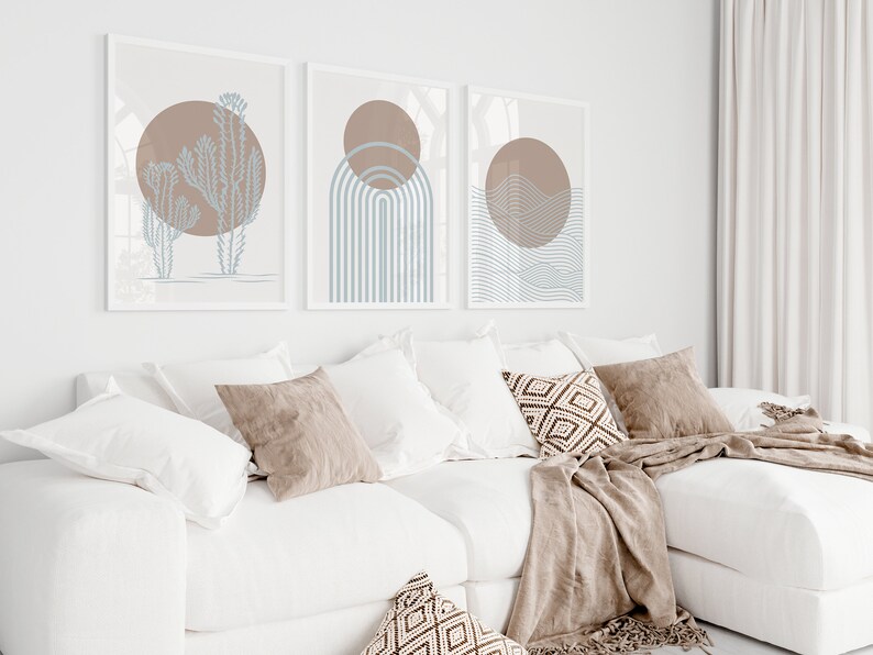 May include: Three abstract art prints with a minimalist design featuring a brown circle and blue lines. The prints are framed in white and hung on a white wall above a white couch with brown throw pillows.