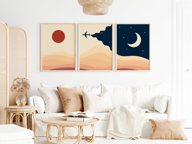 以下が含まれることがあります： 夕焼け、雲の中に飛ぶ飛行機、星のある三日月を描いた抽象的な風景画3点。自然な木製の額縁に収められ、白い壁に掛けられています。白いソファの上にベージュのクッションが置かれています。