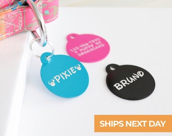 May include: Three round dog tags with personalized text. The tags are blue, pink, and black. The blue tag says "PIXIE" with paw prints. The pink tag has a phone number and says "Puppy at Doggeland". The black tag says "BRUNO".