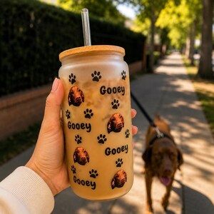 May include: A frosted glass tumbler with a bamboo lid and a clear glass straw. The exterior features a repeating pattern of black paw prints, small photographic portraits of a brown dog, and the word Gooey printed in a black sans-serif font.