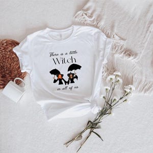 May include: White t-shirt with a black graphic of three witches holding umbrellas. The text reads "There is a little Witch in all of us."