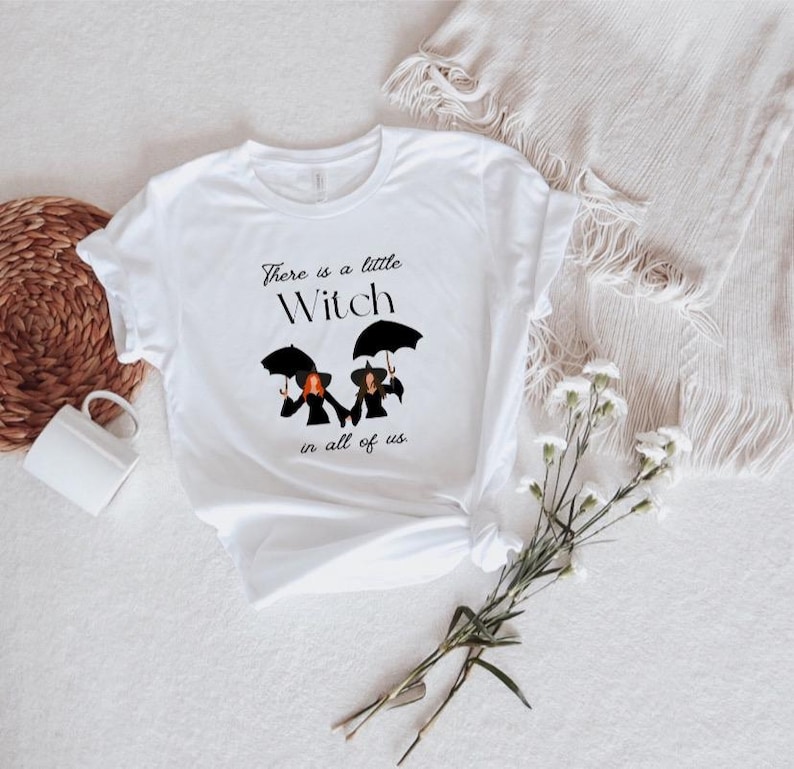 May include: White t-shirt with a black graphic of three witches holding umbrellas. The text reads "There is a little Witch in all of us."
