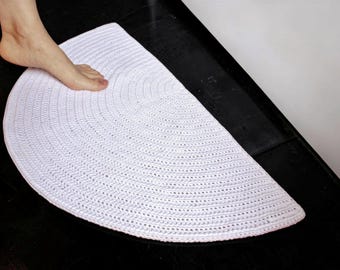 Op de afbeelding: Een witte, halfronde gehaakte badmat. De mat is gemaakt van dik, getextureerd garen en heeft een afgeronde rand. De mat ligt op een zwart oppervlak.