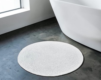 Op de afbeelding: Een witte, ronde, gehaakte badmat met een getextureerd oppervlak. De mat ligt op een grijze betonnen vloer in de buurt van een witte badkuip en een raam. Het ontwerp van de mat heeft concentrische cirkels.