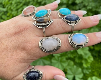 May include: A hand wearing seven silver rings with different colored gemstones. The rings feature a rope-like design around the edges of the oval-shaped stones. The stones are turquoise, rose quartz, tiger's eye, lapis lazuli, moonstone, and two different shades of blue.