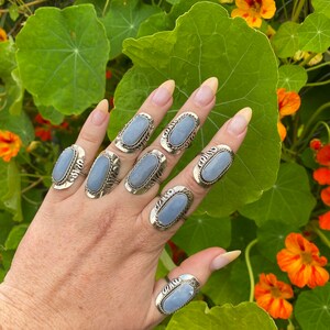 May include: A hand wearing seven silver rings with oval-shaped blue gemstones. The rings are set in a silver band with a decorative design.