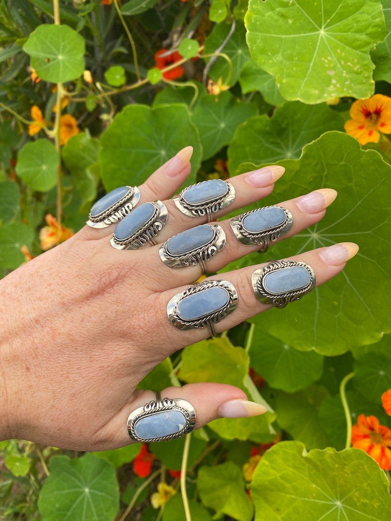 May include: A hand wearing eight silver rings with oval-shaped blue stones. The rings have intricate designs around the stones.
