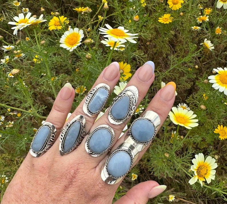 May include: A hand wearing seven silver rings with light blue oval-shaped stones. The rings are set in a silver band with a decorative design.