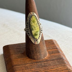 May include: A silver ring featuring an elongated oval-shaped green stone with mottled texture. The stone is set in a smooth silver bezel surrounded by a twisted rope border, mounted on a decorative silver band with openwork scroll details.
