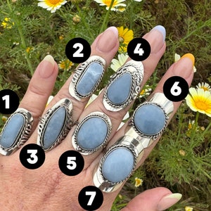 May include: Seven silver rings with blue gemstone settings. The rings are stacked on a hand, with the gemstones facing the camera. The rings are all different shapes and sizes.