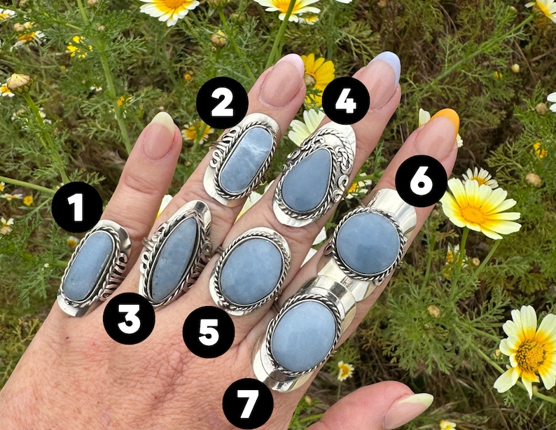 May include: Seven silver rings with blue gemstone settings. The rings are stacked on a hand, with the gemstones facing the camera. The rings are all different shapes and sizes.