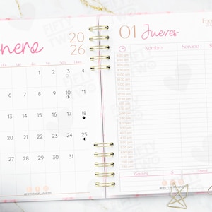 Puede incluir: Agenda abierto con un diseño rosa y blanco. La página izquierda muestra un calendario para enero de 2026, mientras que la página derecha es para programar citas. El planificador tiene una encuadernación en espiral dorada y un lápiz rosa.