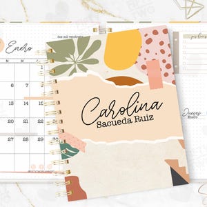 May include: A spiral-bound planner with a cover featuring the name "Carolina" in elegant script. The design includes abstract shapes in muted colours, a calendar, and the text "Fifty Two Planners".