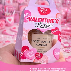 Puede incluir: Una caja de regalo rosa de San Valentín con asa, que contiene un pequeño frasco de gel de manos Cozy Vanilla Almond. La caja tiene detalles en forma de corazón y las palabras "Valentine's Day". El frasco es visible a través de un recorte en forma de corazón.